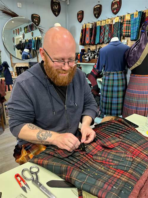 A bearded man wearing glasses is shown pinning a tartan kilt fabric on a table inside a shop. The kilt features a classic red, green, and blue plaid pattern.